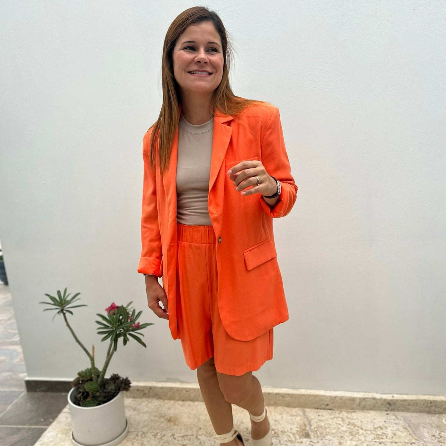 A women wearing a orange color blazer, beige top and orange shorts.