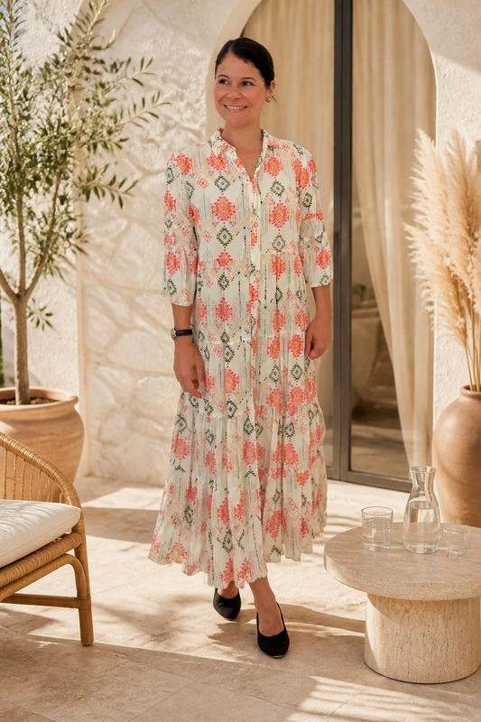 Woman wearing ivory cotton maxi dress with coral and sage aztec geometric print, 3/4 sleeves, tiered skirt and button-front collar, styled with black flat mules in a Mediterranean terrace setting
