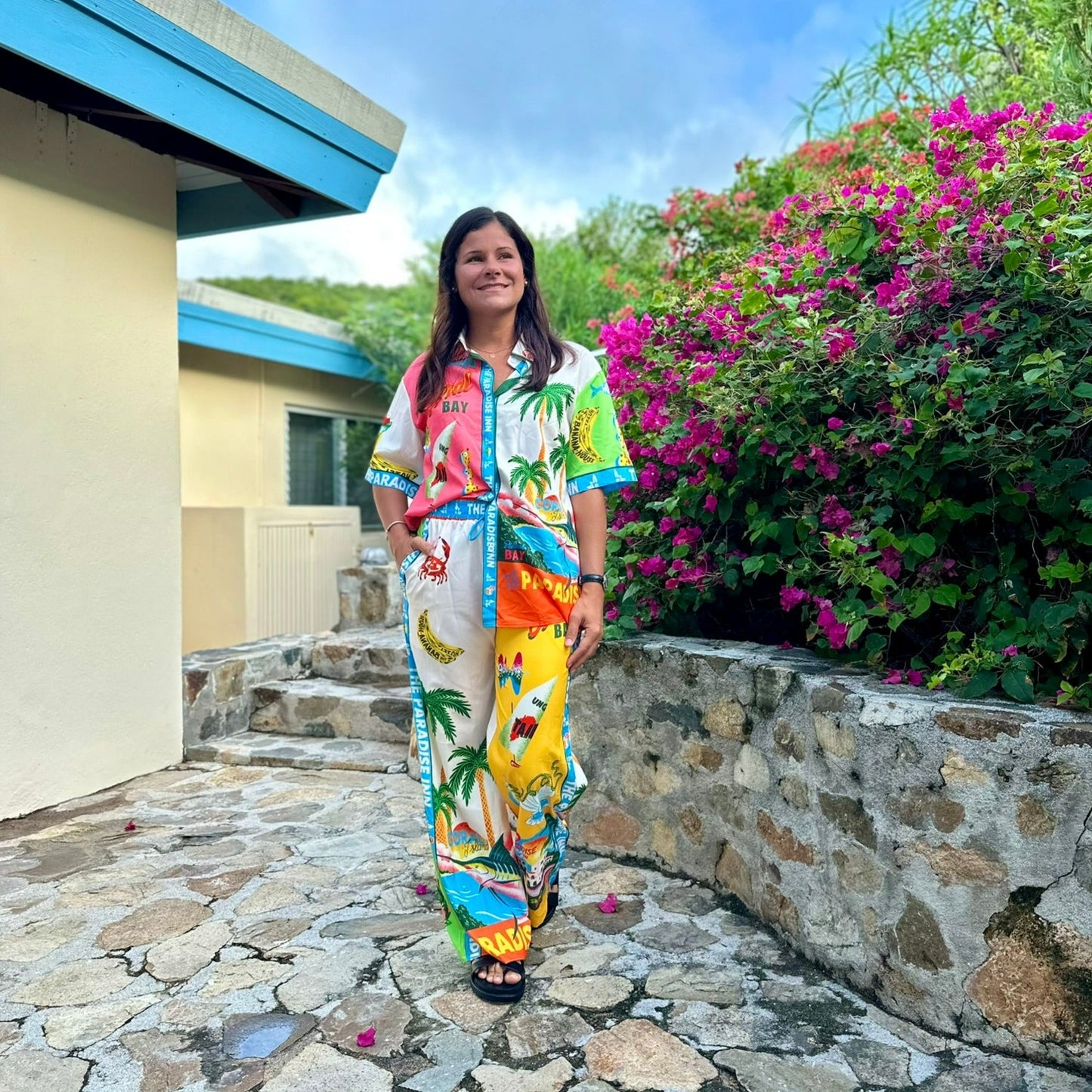 Woman wearing colorful tropical print two-piece set with palm trees, bananas, and beach graphics in pink, yellow, blue, and orange, standing outdoors with flowering bougainvillea in background