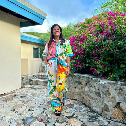 Woman wearing colorful tropical print two-piece set with palm trees, bananas, and beach graphics in pink, yellow, blue, and orange, standing outdoors with flowering bougainvillea in background