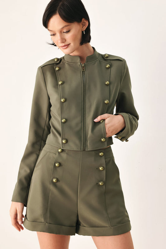 Woman wearing structured olive green cropped military jacket with double-breasted antique gold buttons, mock neckline and front zipper closure, styled with matching olive shorts and black heels