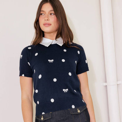 Woman wearing a navy polka dot top with a white collar and a dark denim skirt against a white background.