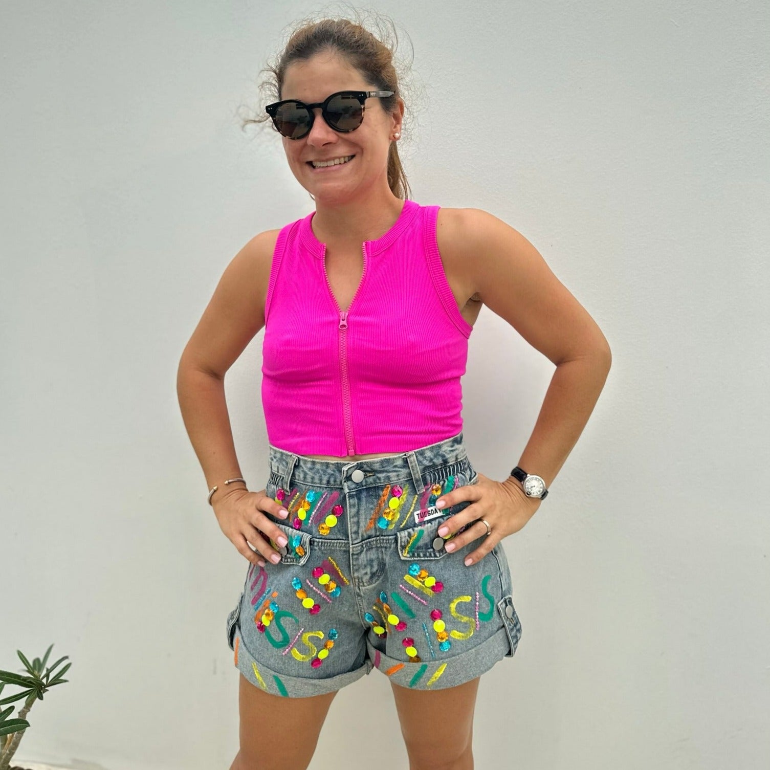 A person wearing hand painted denim shorts with a high-rise waist and colorful bead details, paired with a pink sleeveless top.
