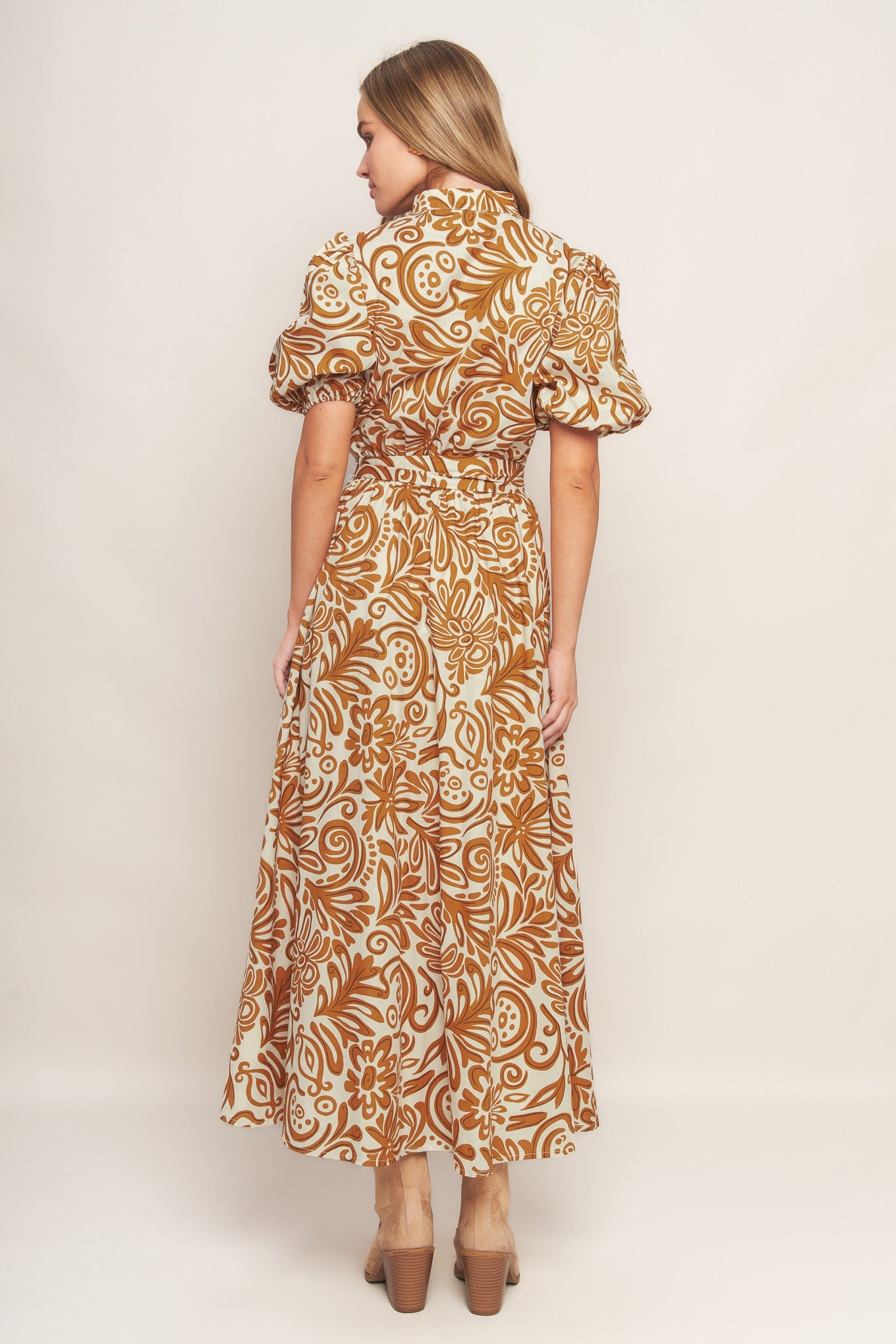 Woman wearing a camel color floral patterned maxi dress with puffed sleeves against a plain background, presnting the back of the dress