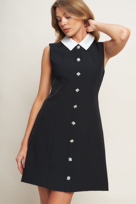 Woman wearing a black sleeveless dress with white collar and buttons on a plain background