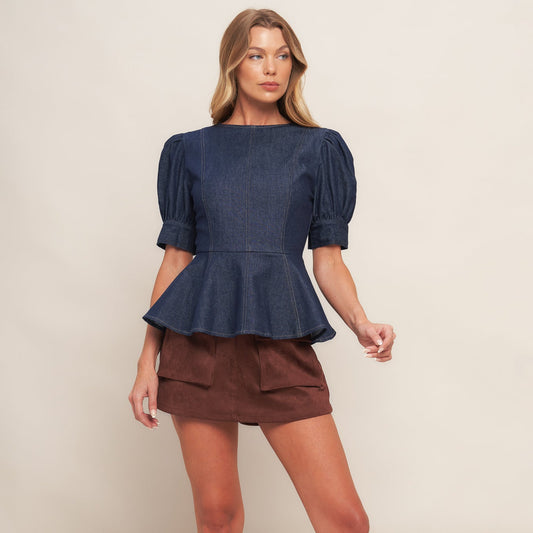 Woman wearing a navy peplum top and brown skirt with black boots on a beige background