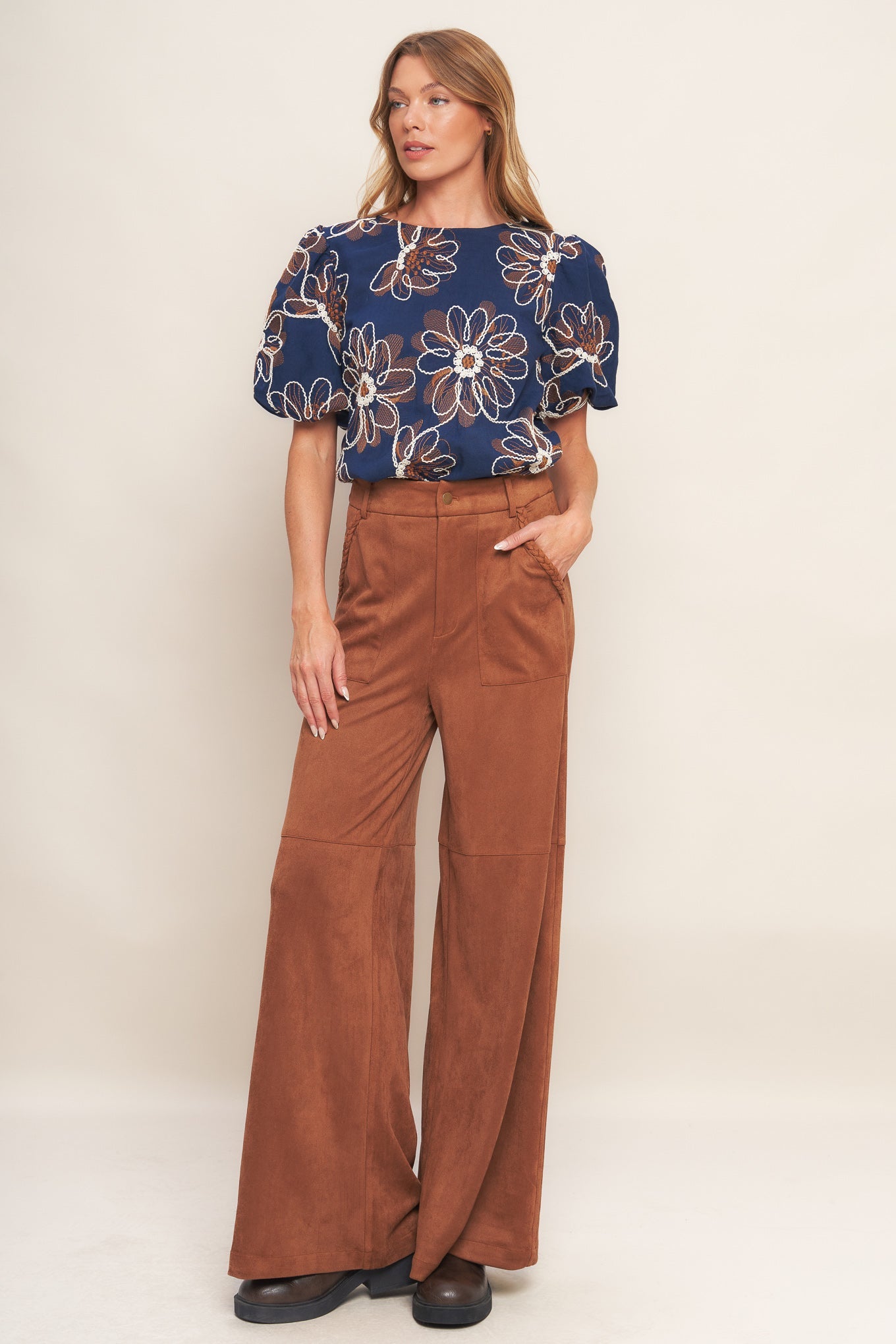 Woman wearing a blue floral top and brown flared pants on a white background