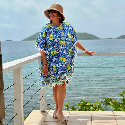 A women wearing a blue kaftan with a lemon print, standing on a balcony by the sea.
