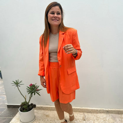 A women wearing a orange color blazer, beige top and orange shorts. 