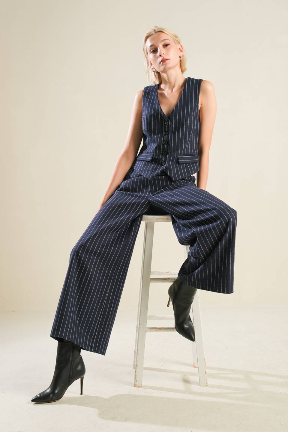 Woman wearing a blue pinstripe jumpsuit sitting on a stool against a plain background