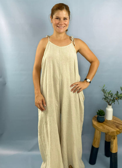 A female model wearing a beige linen jumpsuit with a sleeveless top and maxi length pants, standing indoors.