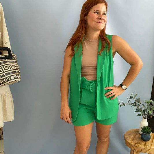 A women wearing a green color vest, beige top and green belted shorts. 